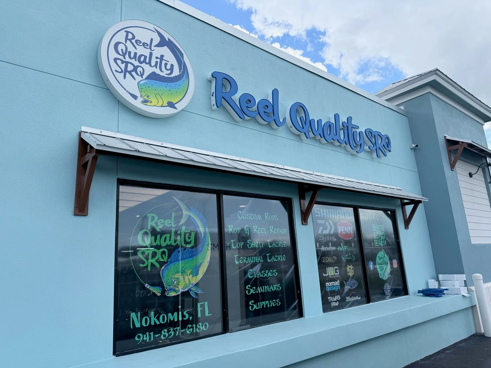 Light-blue Reel Quality SRQ storefront with large blue signage, the circular mahi-mahi logo, and colorful fish artwork on the windows.